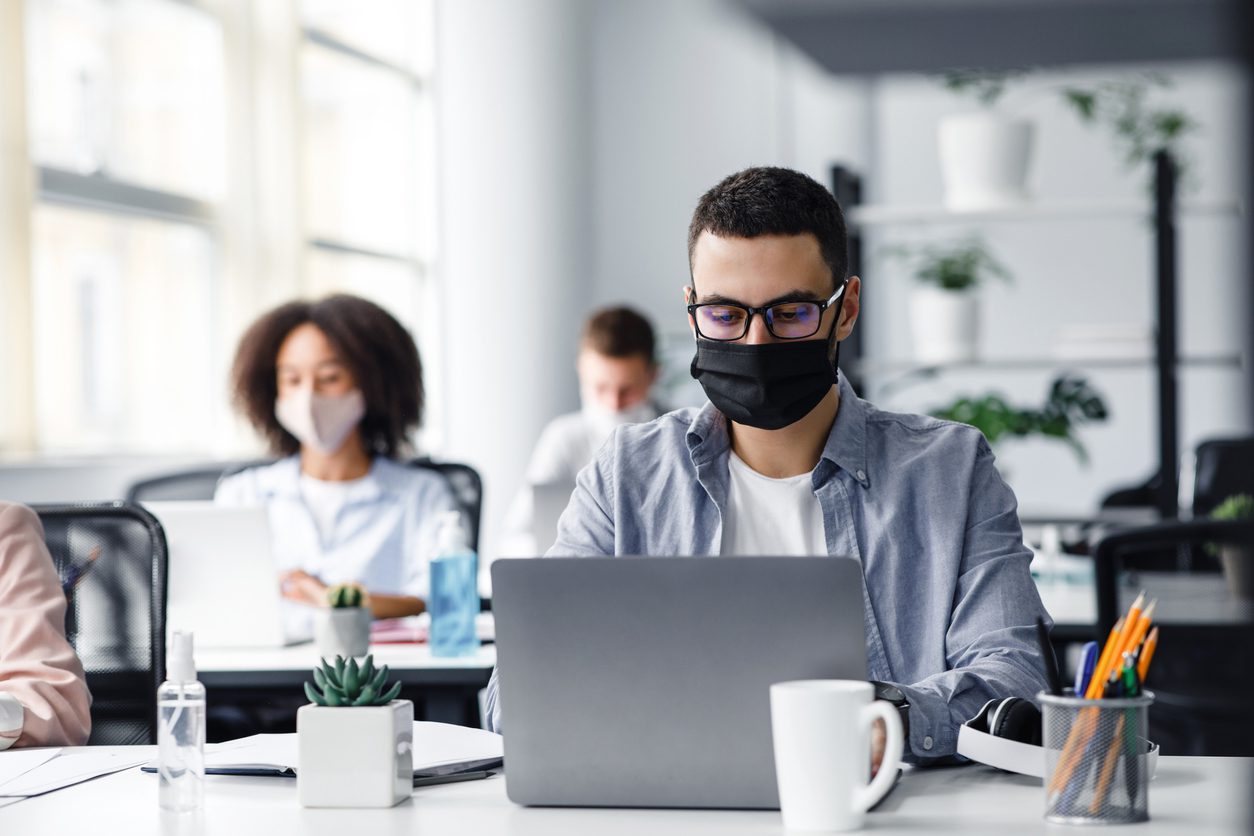Office center workers are protected from virus outbreak during covid-19 epidemic. Young hipster man in glasses and protective mask works at laptop