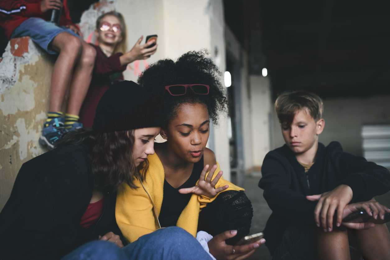 Group of teenagers gang sitting indoors in abandoned building, using smartphones.