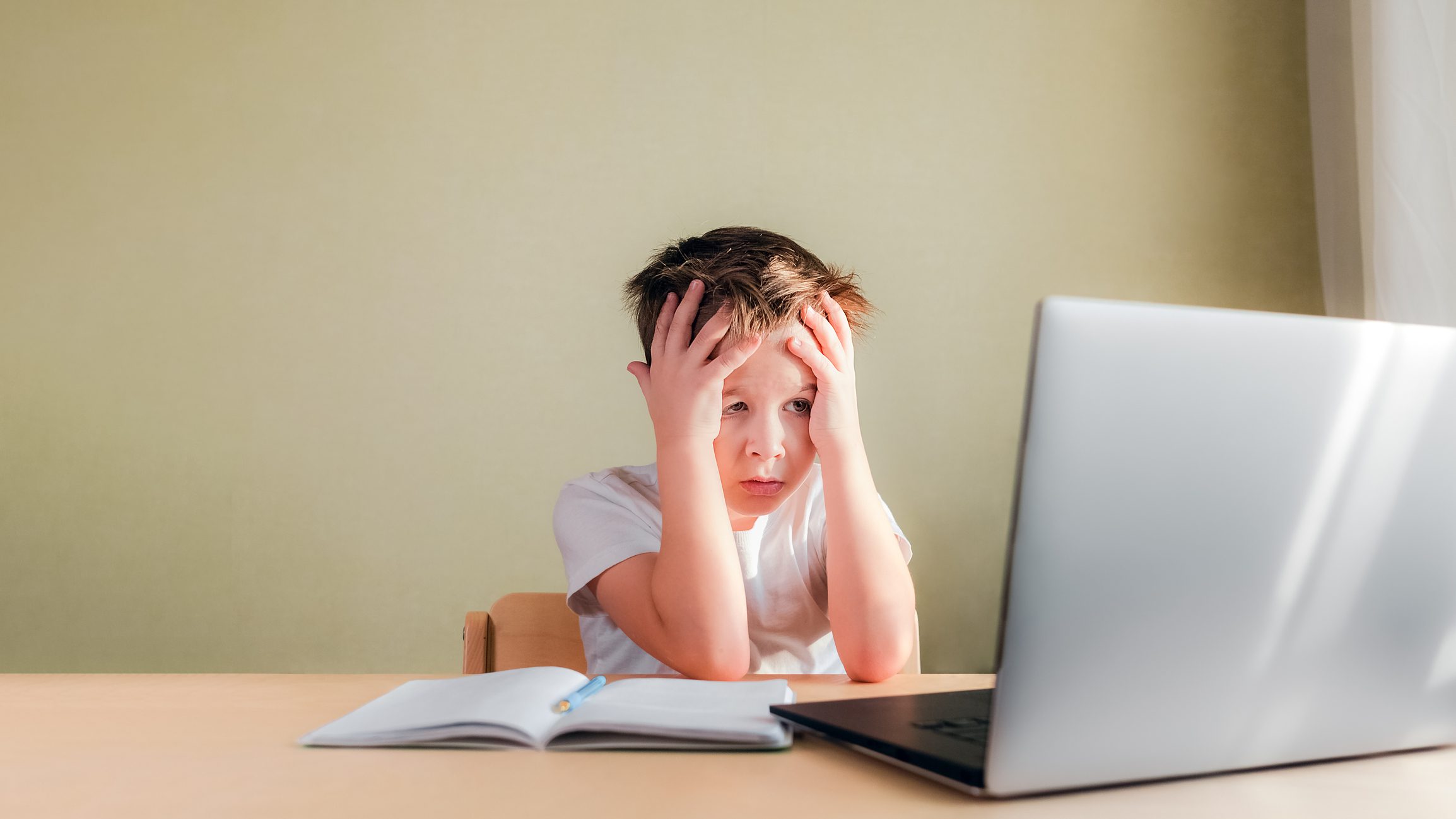 El niño se sienta en el escritorio, mira tristemente la computadora y sostiene su cabeza con las manos